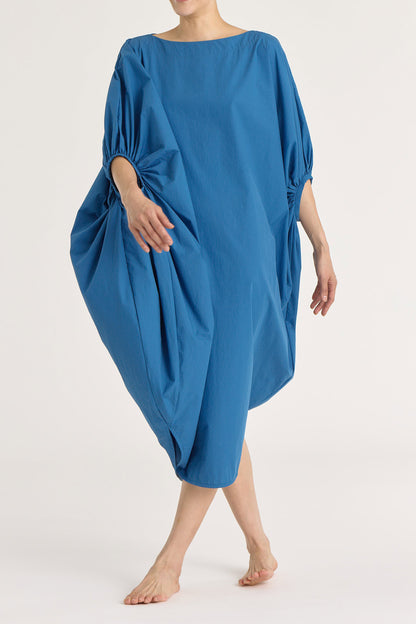 A person wearing a blue teardrop dress with a wide boat neck and voluminous sleeves, loose-fitting cotton dress.