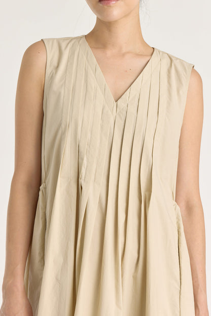 A model wearing a beige pleated V-neck dress with side seam pockets and an adjustable drawstring at the back.