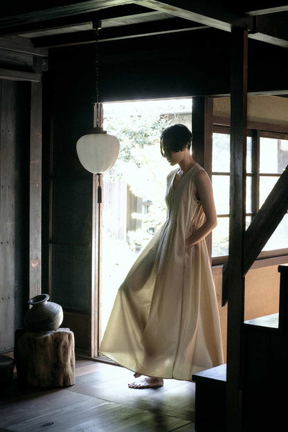 A woman wearing a pleated V-neck dress in an oyster color, standing in a room with wooden elements and a dark color scheme.