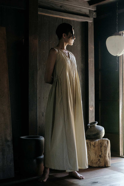 A woman wearing a pleated V-neck dress in an oyster color, standing in a room with wooden elements and a dark color scheme.