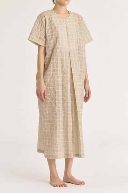 A beige crew neck dress with a single button closure at the front, inverted pleat, and side seam pockets. The dress appears to be made of a lightweight, slightly sheer fabric and is shown being worn by a model.