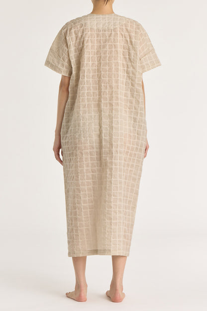 A beige crew neck dress with a single button closure at the front, inverted pleat, and side seam pockets. The dress appears to be made of a lightweight, slightly sheer fabric and is shown being worn by a model.