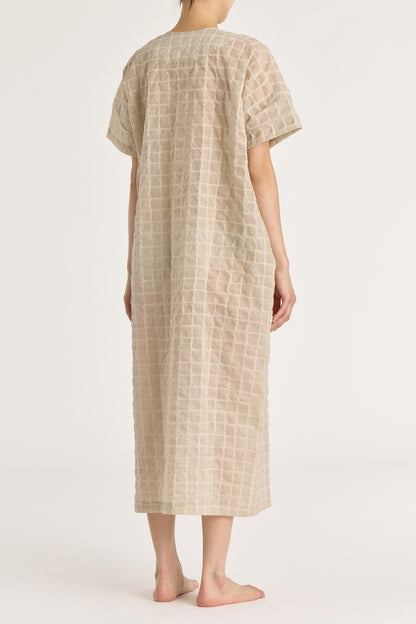 A beige crew neck dress with a single button closure at the front, inverted pleat, and side seam pockets. The dress appears to be made of a lightweight, slightly sheer fabric and is shown being worn by a model.