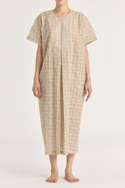 A beige crew neck dress with a single button closure at the front, inverted pleat, and side seam pockets. The dress appears to be made of a lightweight, slightly sheer fabric and is shown being worn by a model.