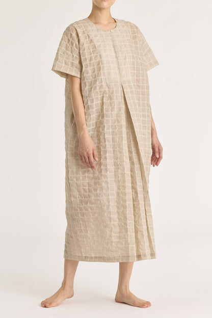A beige crew neck dress with a single button closure at the front, inverted pleat, and side seam pockets. The dress appears to be made of a lightweight, slightly sheer fabric and is shown being worn by a model.