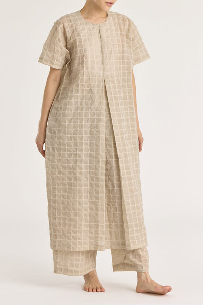 A beige crew neck dress with a single button closure at the front, inverted pleat, and side seam pockets. The dress appears to be made of a lightweight, slightly sheer fabric and is shown being worn by a model.