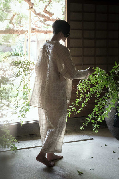 A person standing by a window wearing a beige, long, sheer shirt with a relaxed fit.