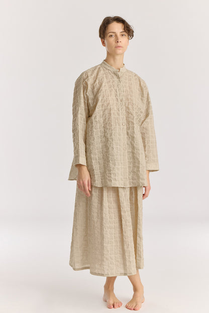 A person standing wearing a beige, long-sleeved, stand-collar shirt with button closures and a matching long skirt.